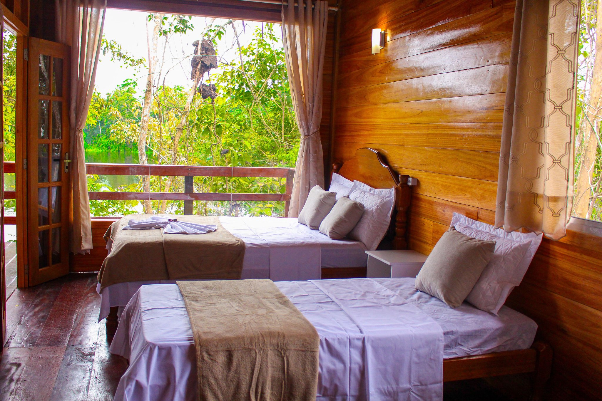 Rustic hardwood walls, fresh linens, and the rainforest right outside your door. A twin-bed jungle lodge room with warm timber walls, white linens, and an open balcony door framing a lush rainforest waterway — part of a manaus jungle tour package