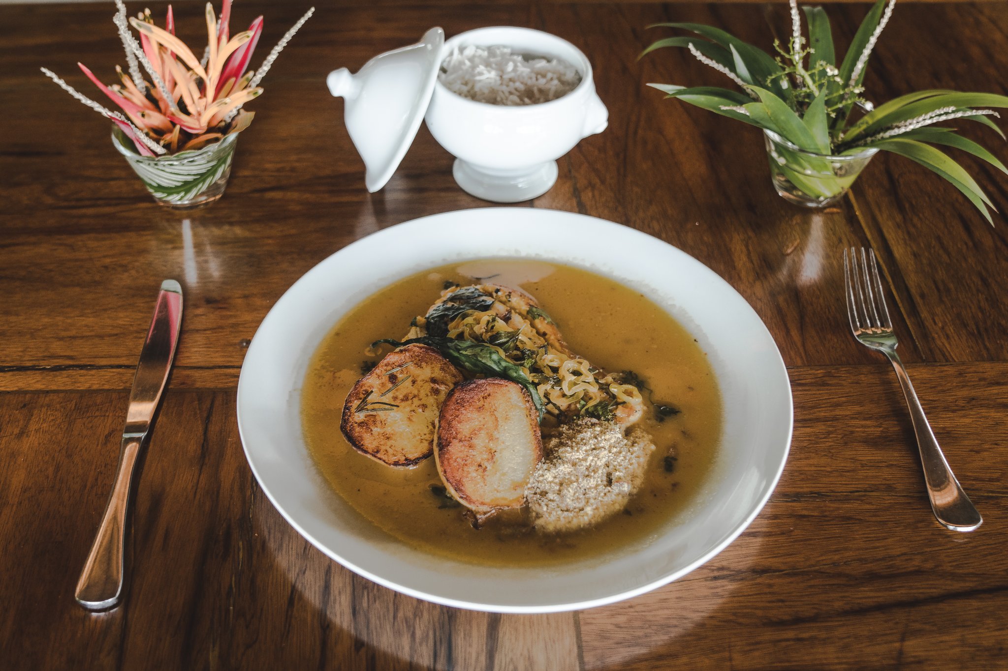 Uma tigela branca com caldo amazônico, medalhões dourados, folhas verdes e farinha de mandioca servida no restaurante do lodge Mirante do Gavião sobre uma mesa de madeira polida, com uma sopeira branca de arroz e arranjos florais tropicais