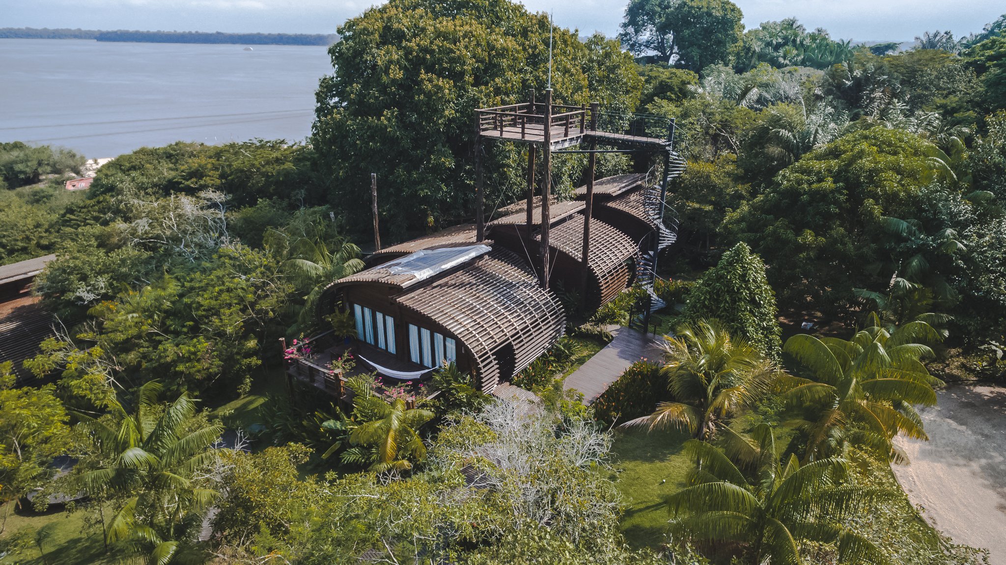 Vista aérea do Mirante do Gavião Lodge com sua arquitetura curvilínea em madeira integrada à densa floresta amazônica às margens do Rio Negro, com passarela elevada e torre de observação acima das copas das árvores