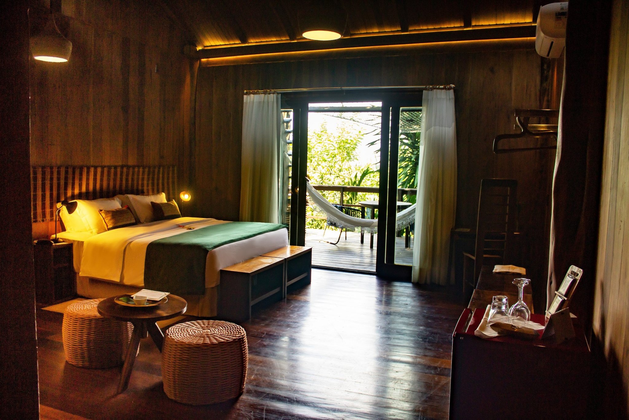Jungle comfort awaits — your private balcony and hammock are just steps from the bed. Warmly lit Mirante do Gavião lodge bedroom with a double bed, wicker ottomans, and open French doors revealing a private balcony with a hammock overlooking the Amazon rainforest canopy