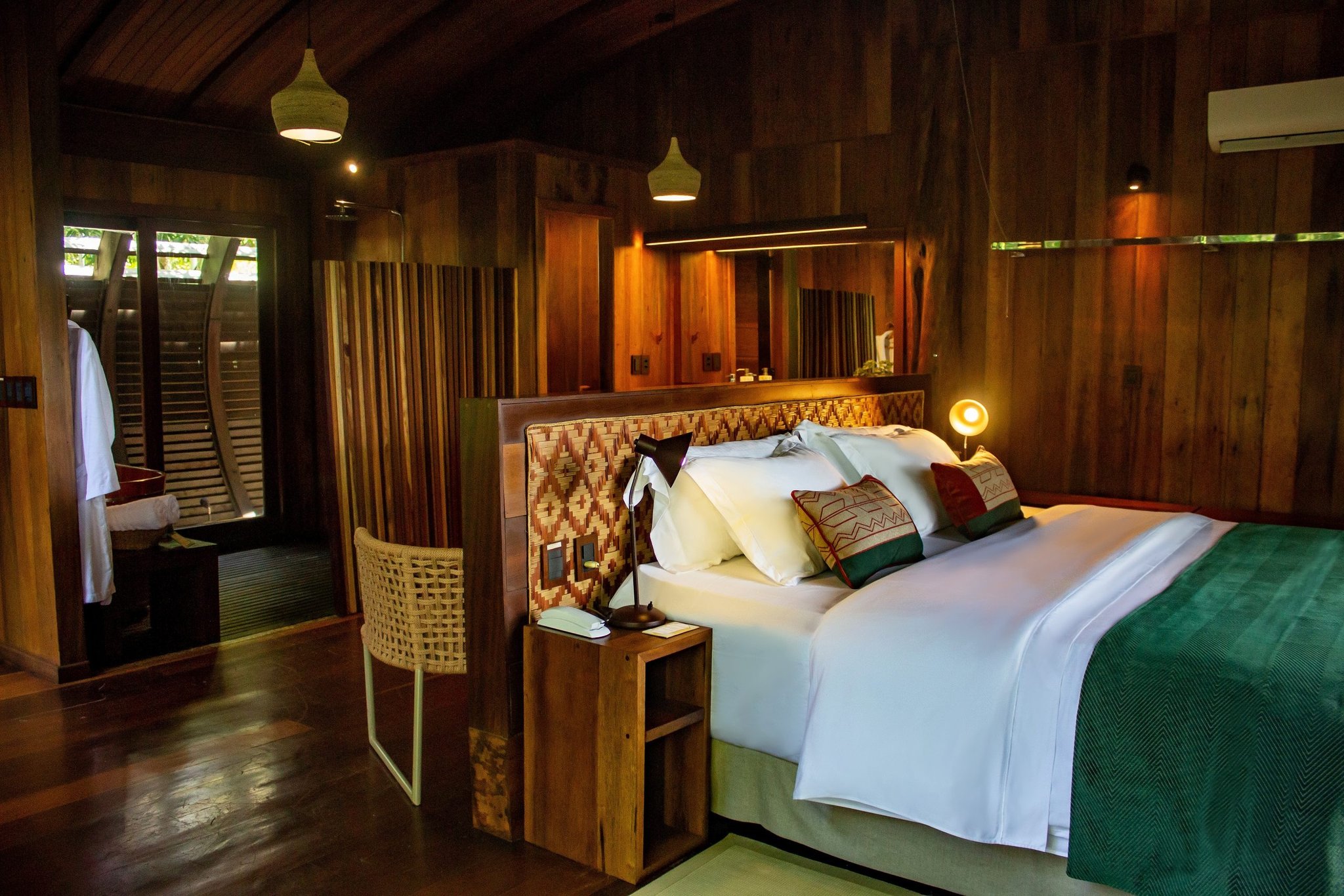 Quarto iluminado do lodge Mirante do Gavião com cama king-size em linho branco, almofadas com estampas amazônicas coloridas, cabeceira em rattan trançado e paredes de madeira escura