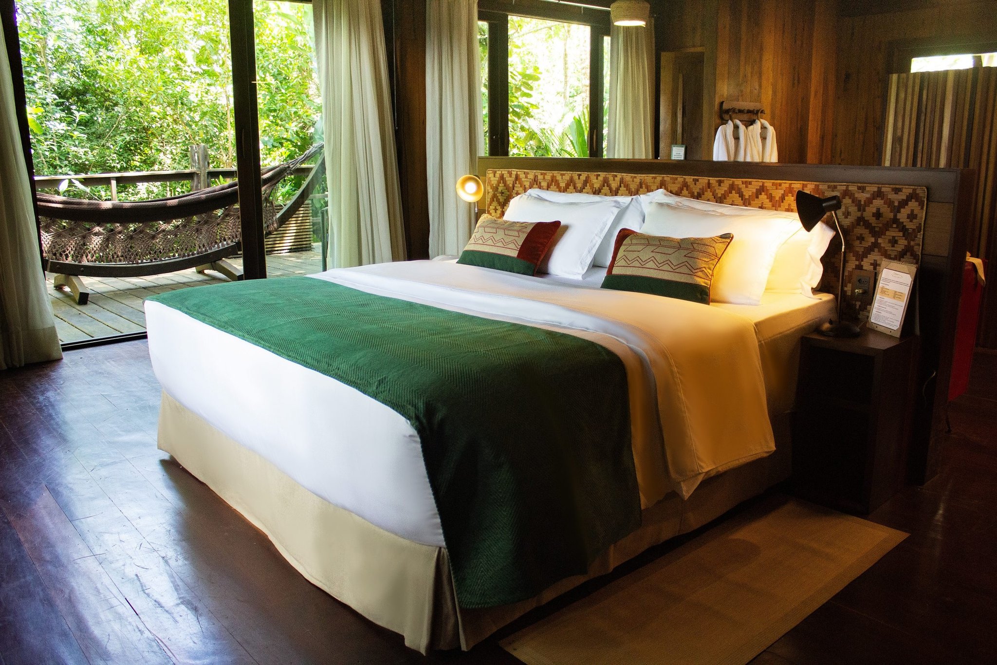 Cama king size com pé-de-cama verde e cabeceira trançada em um bangalô da floresta no Mirante do Gavião, varanda de madeira com rede e copa da Amazônia visível pelas portas de vidro