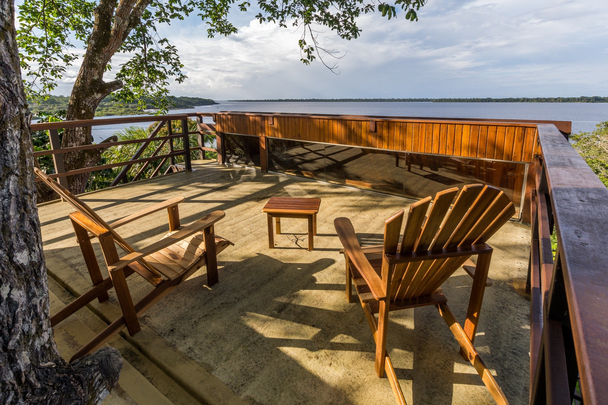 Duas cadeiras de madeira e uma mesinha no deck aberto do Mirante do Gavião, com vista panorâmica para o Rio Negro e a floresta amazônica ao fundo