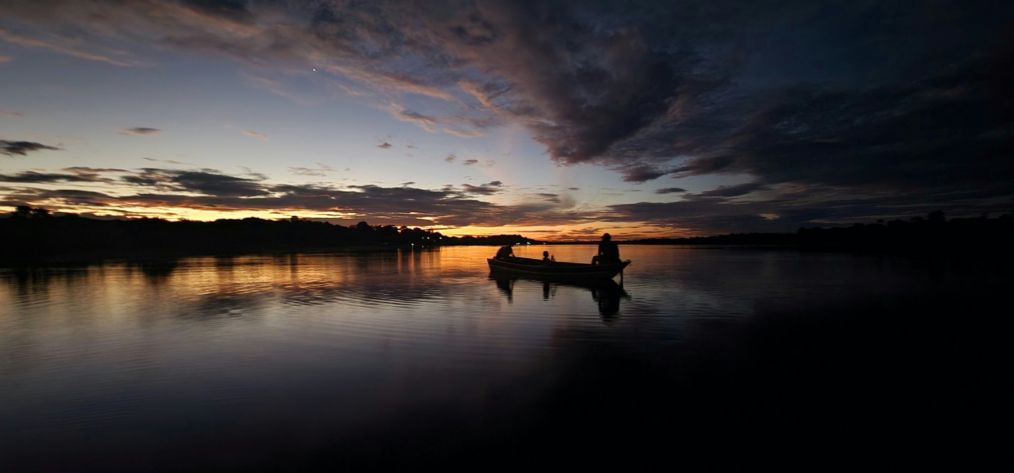 Silhueta de três pessoas em um pequeno barco de madeira no Rio Negro ao entardecer, com o pôr do sol laranja e dourado refletido na água calma próximo ao Mirante do Gavião