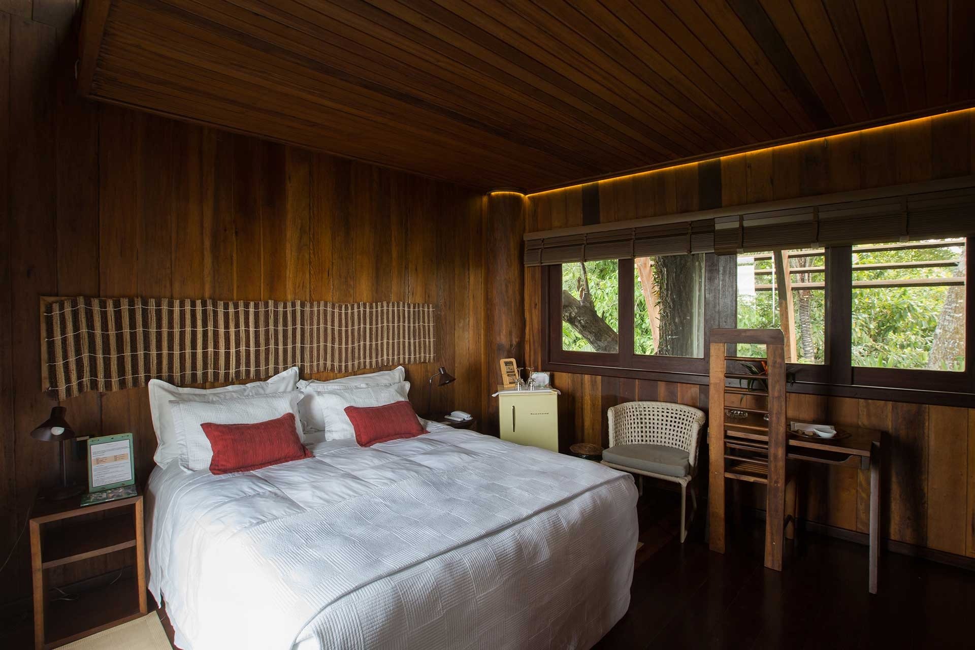 Quarto com paredes de madeira escura no Mirante do Gavião, cama de casal com roupa de cama branca, almofadas vermelhas e janelas com vista para a floresta amazônica