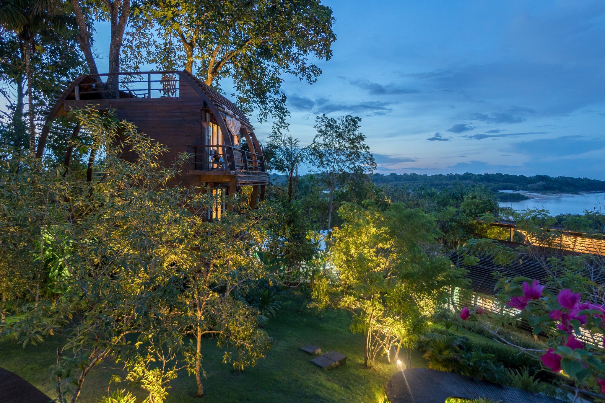 Uma cabana de madeira arqueada do Mirante do Gavião brilha com luz quente ao entardecer, cercada por jardins exuberantes da floresta amazônica com vista para o Rio Negro