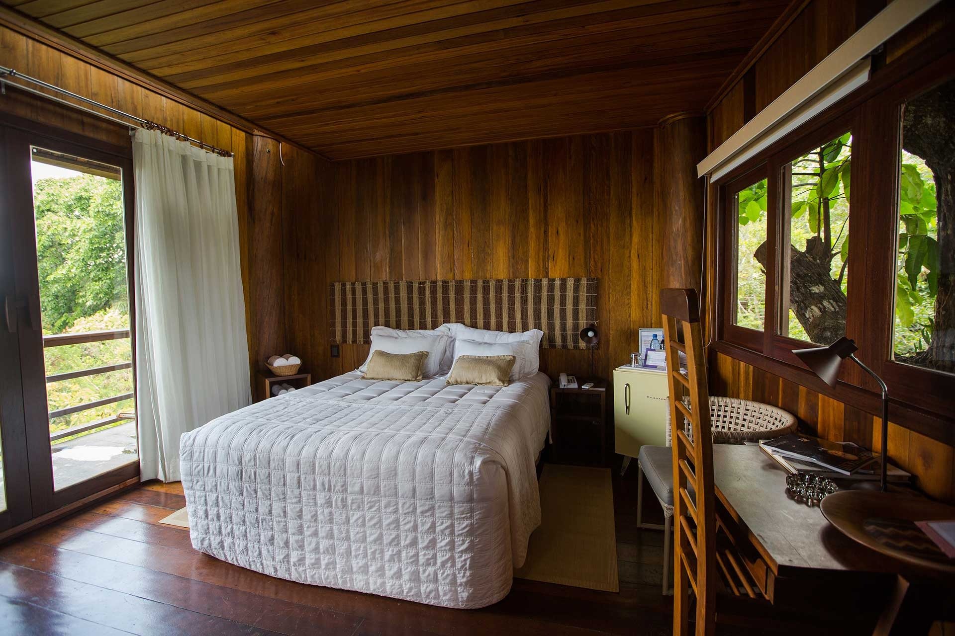 Quarto de casal aconchegante no Mirante do Gavião com paredes revestidas de madeira, colcha branca acolchoada, cabeceira trançada e porta de vidro aberta para a floresta amazônica