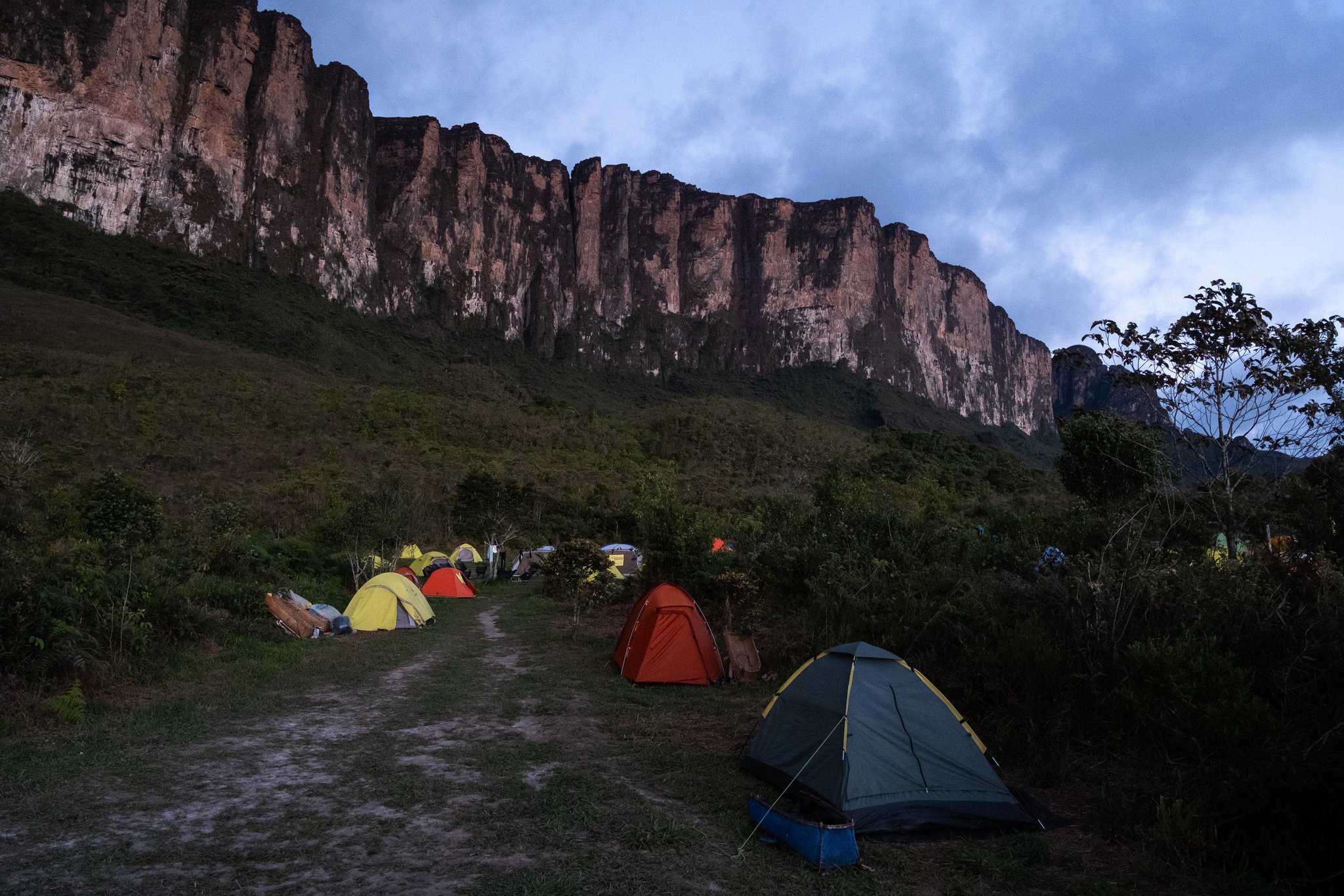 Colorful dome tents pitched on a grassy slope during the Mount Roraima hike, with the towering ancient sandstone cliffs of the tepui rising dramatically under a moody dusk sky