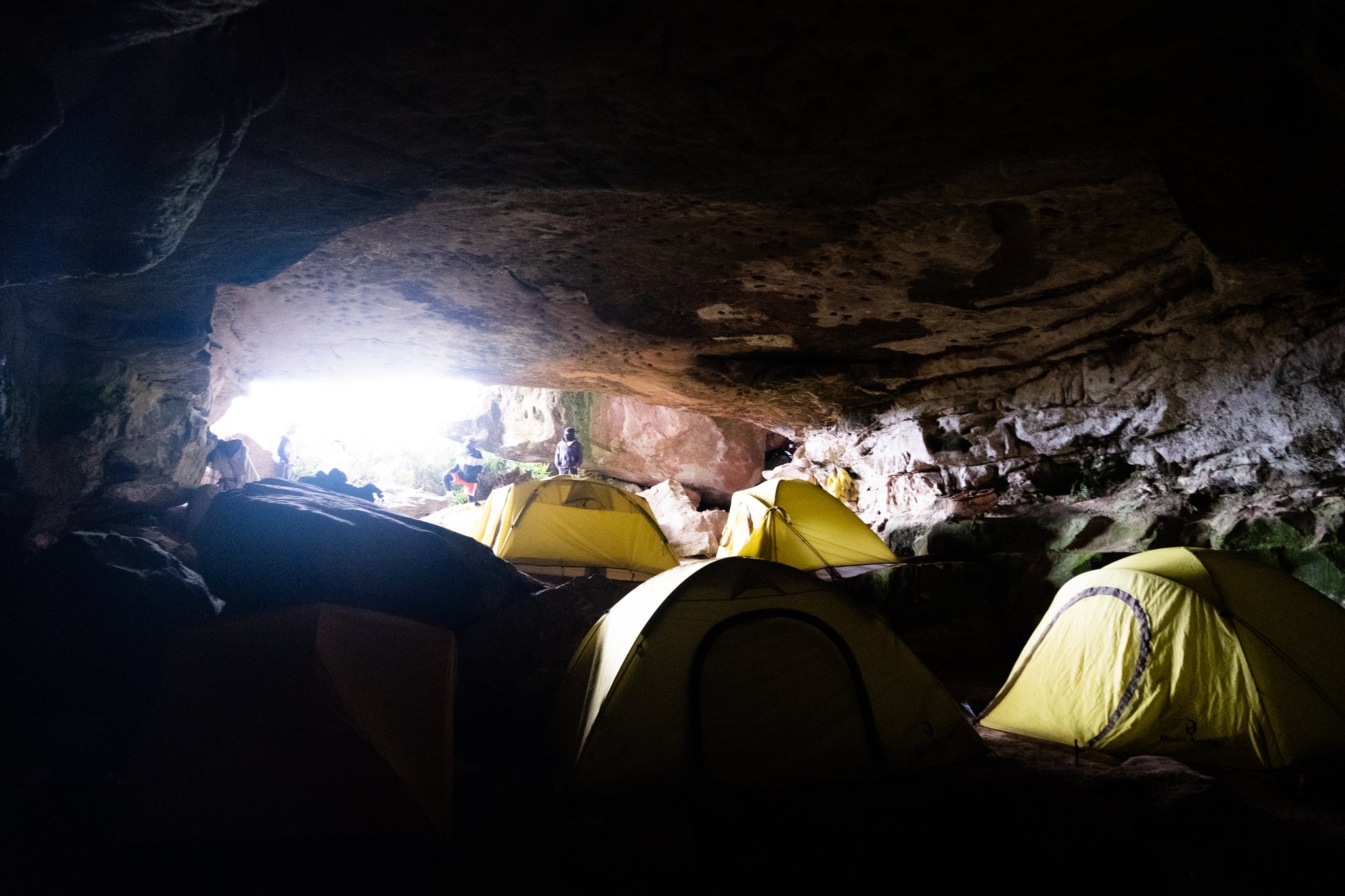 Yellow dome tents pitched inside a rocky cave shelter on the Mount Roraima hike, with daylight streaming through a low entrance where hikers stand silhouetted