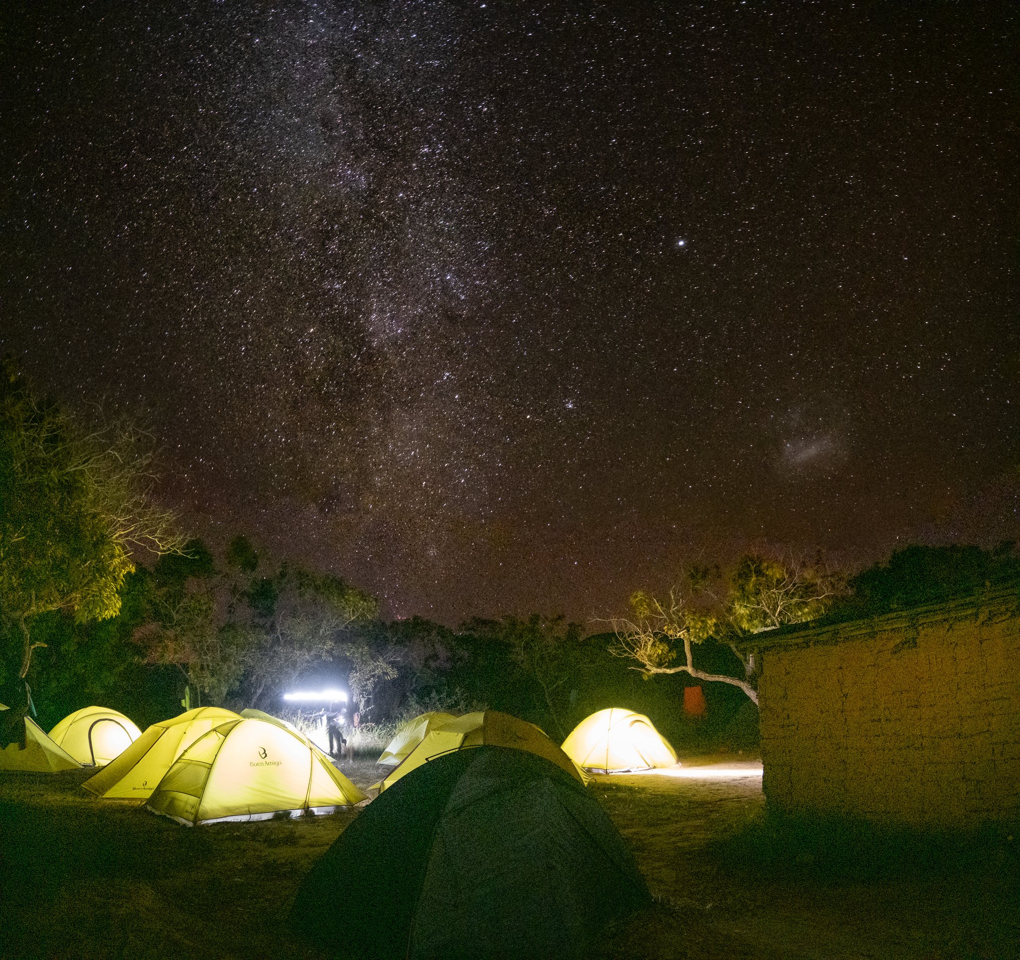 Glowing yellow tents clustered on the Mount Roraima hike beneath a vast Milky Way sky, with a lone figure standing in the camp light and sparse trees silhouetted on the horizon