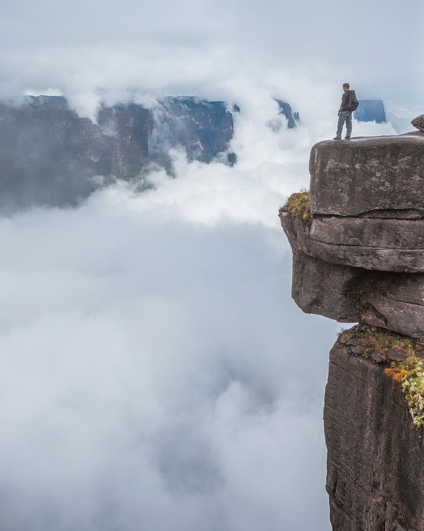 A lone hiker with a backpack stands on the edge of an ancient sandstone rock pillar high above a sea of clouds on a Mount Roraima hike, with a massive tepui escarpment emerging from the mist in the distance