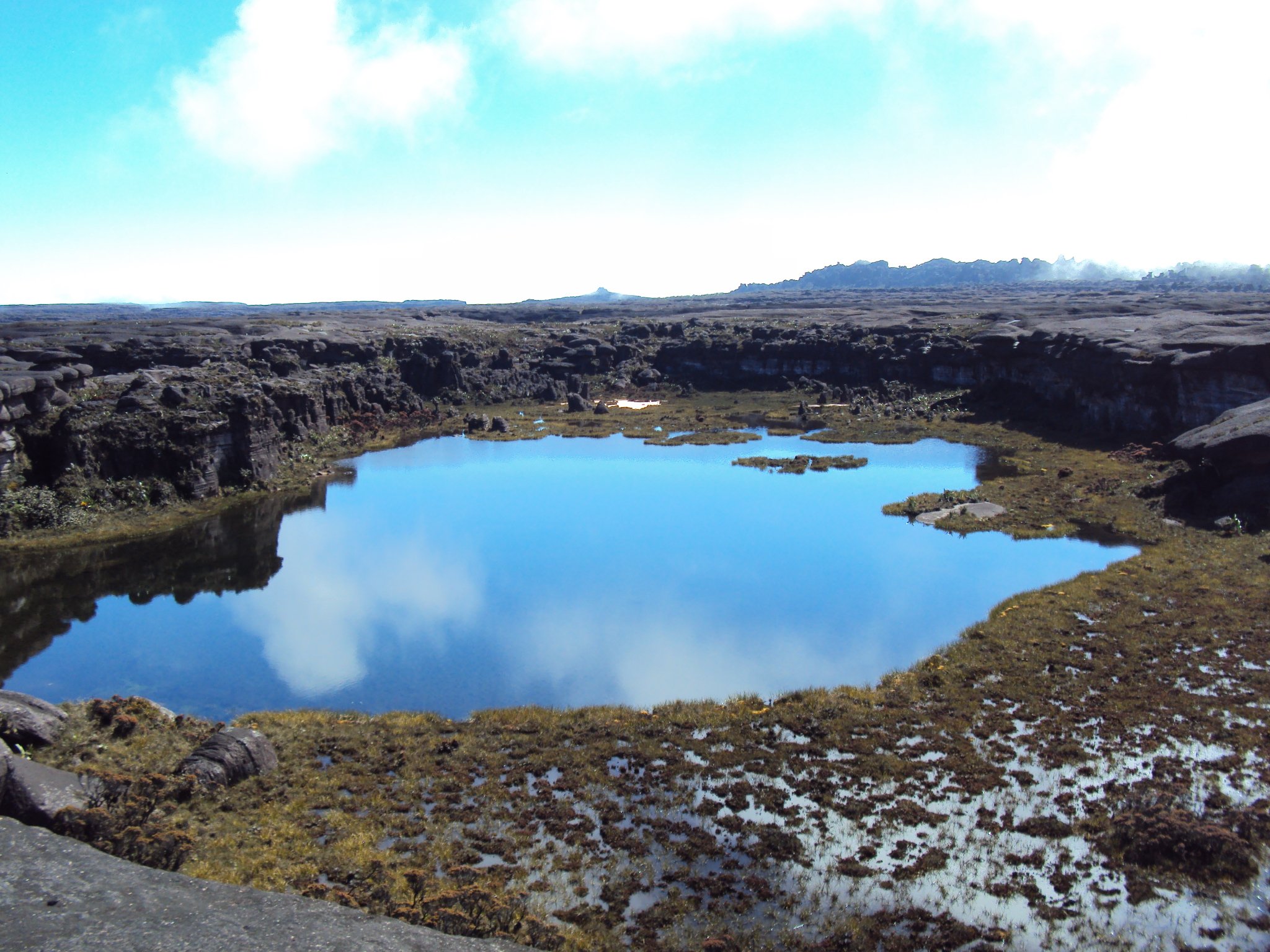 A still blue lagoon reflects the sky on the ancient rocky summit plateau of the Mount Roraima hike, surrounded by dark weathered sandstone cliffs and golden-brown tepui vegetation