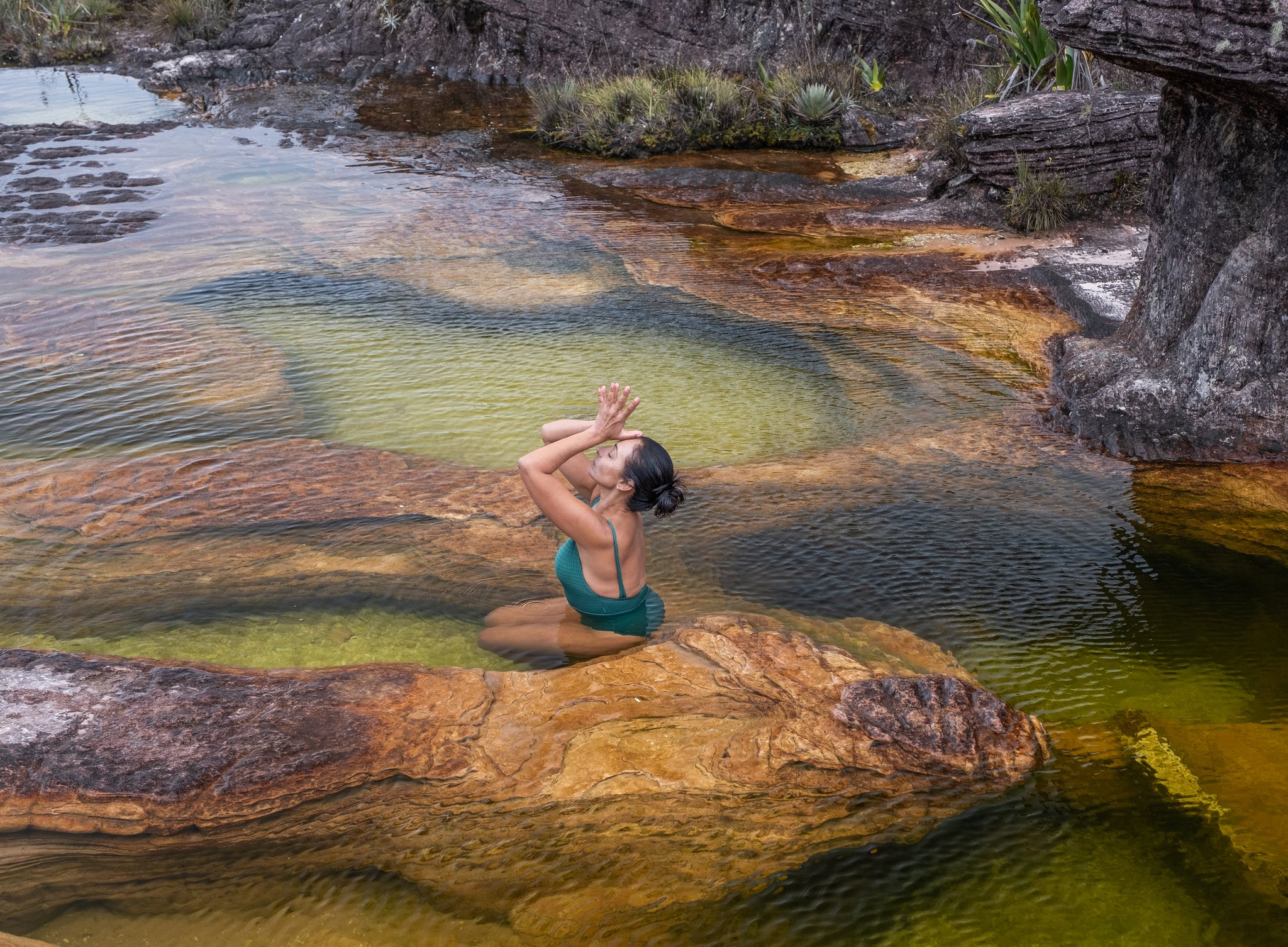 A woman in a teal swimsuit kneels meditatively in a shallow natural pool on the Mount Roraima hike, surrounded by rust-coloured layered rock and crystal-clear green water