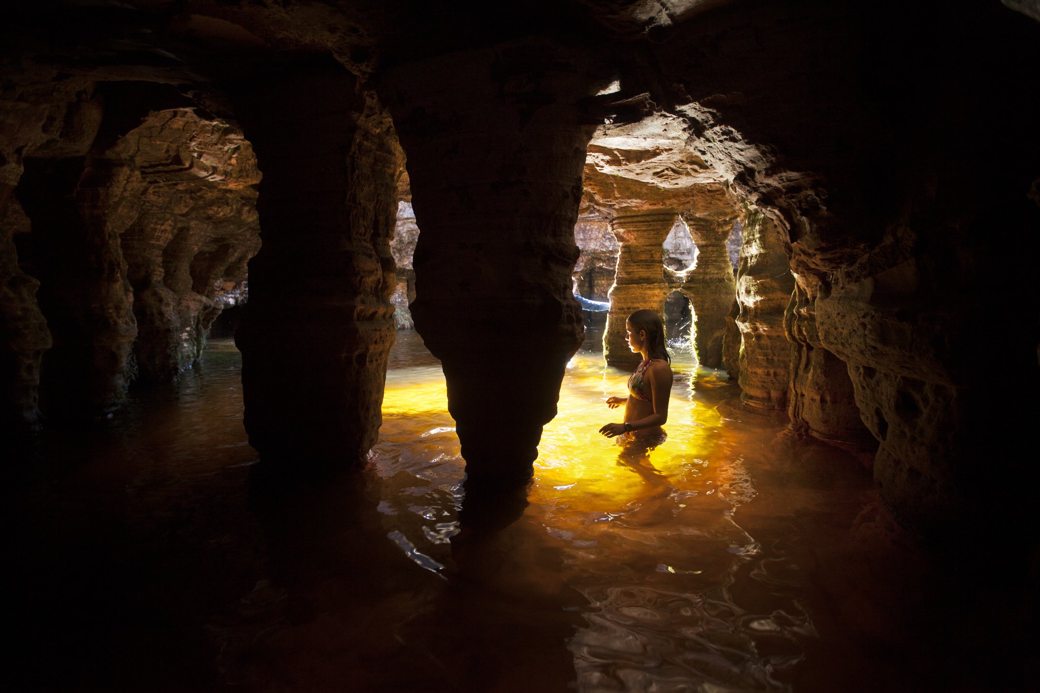 A woman wading waist-deep through a glowing amber pool inside an ancient sandstone cave chamber, lit by warm golden light filtering through eroded rock arches on the Mount Roraima hike