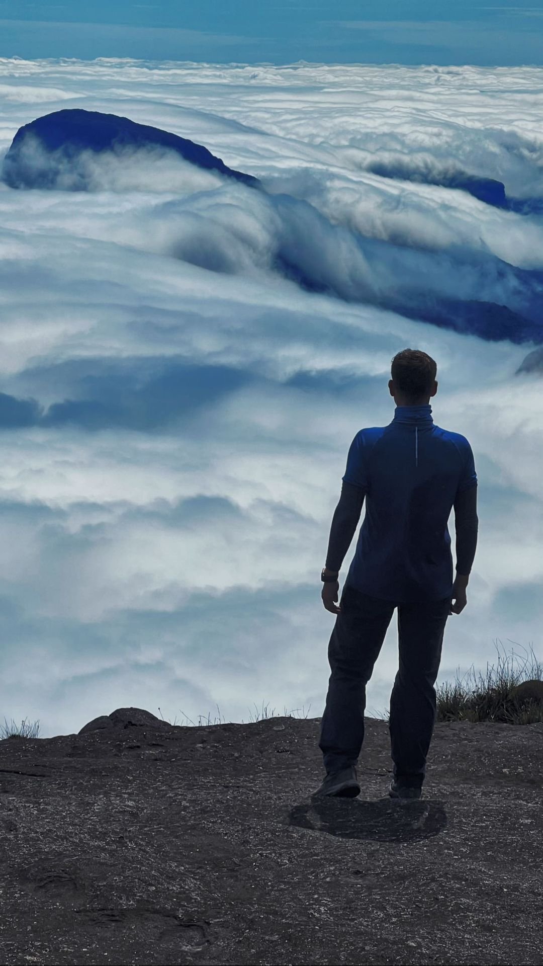 A hiker stands on a rocky summit ledge of the Mount Roraima hike, gazing out over a vast billowing sea of clouds with dark tepui peaks rising through the white mass below