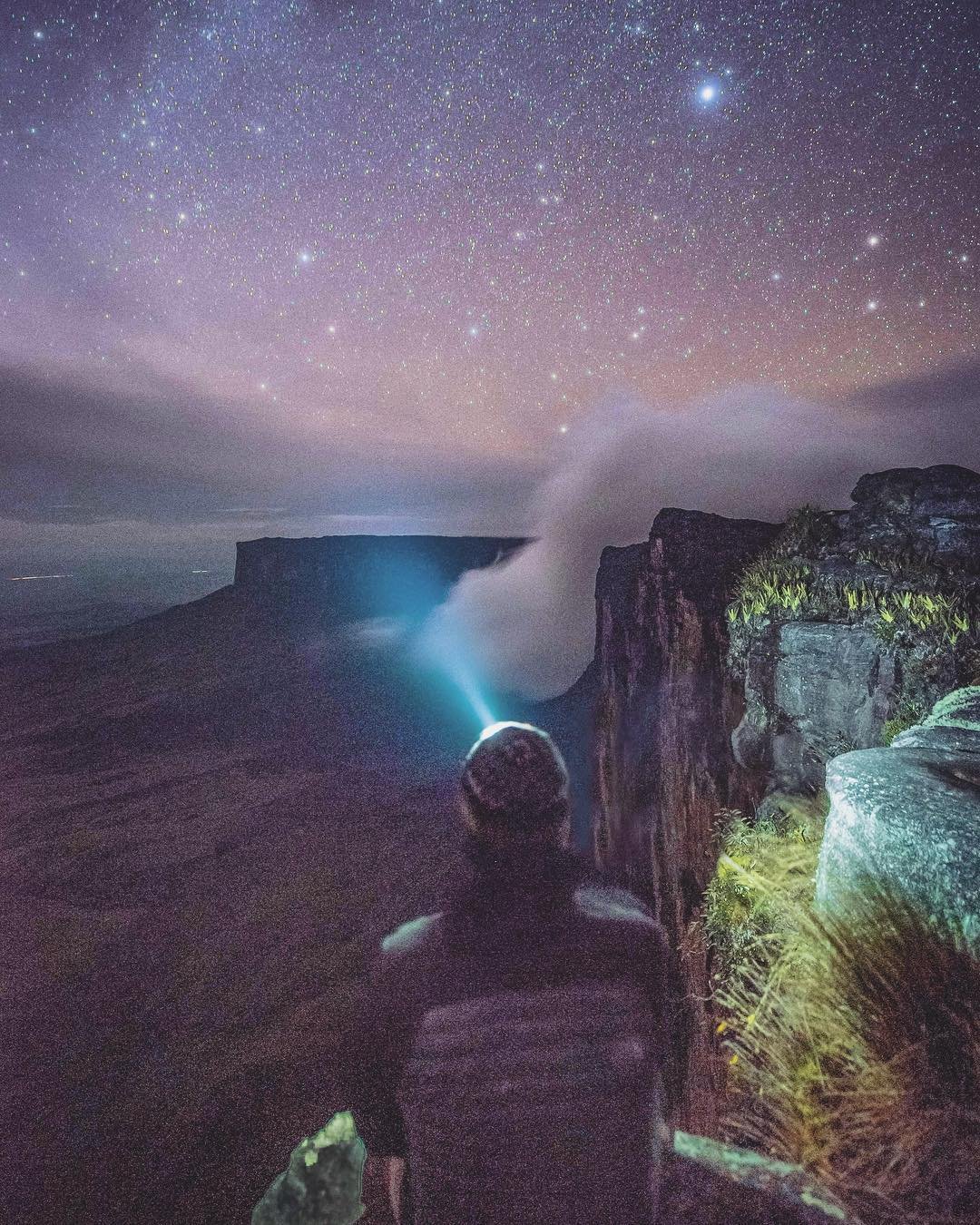 A lone hiker with a headlamp stands on the rocky summit plateau of a tepui at night, gazing toward a flat-topped cliff shrouded in mist beneath a vast star-filled sky on the Mount Roraima hike