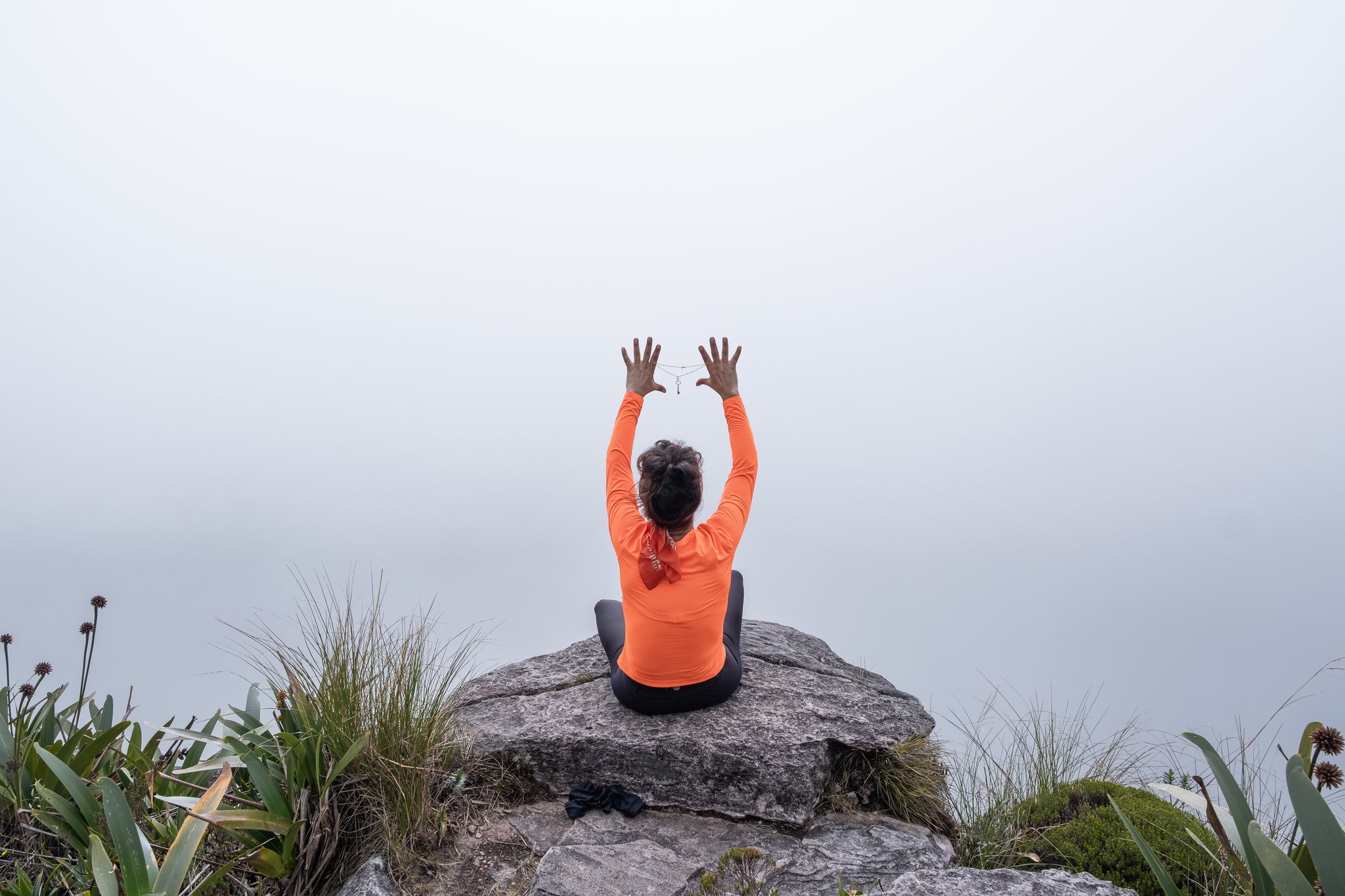 A person in an orange long-sleeve top sits cross-legged on a rocky summit outcrop, arms raised toward a dense white fog, surrounded by high-altitude bromeliads and grasses during the Mount Roraima hike