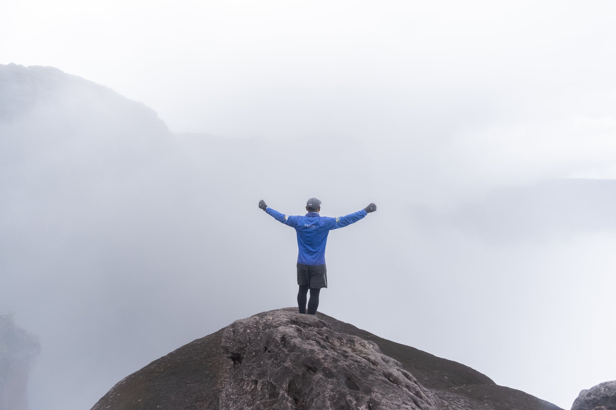A hiker in a blue jacket stands arms outstretched on a rocky summit outcrop during the Mount Roraima hike, surrounded by thick cloud and mist