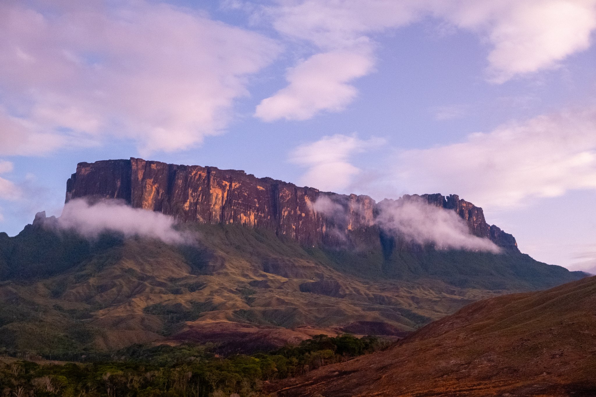 The flat-topped sandstone cliff of a tepui glows amber in warm light during the Mount Roraima hike, with low clouds wrapping its base and rolling green highlands stretching below under a violet-tinged sky