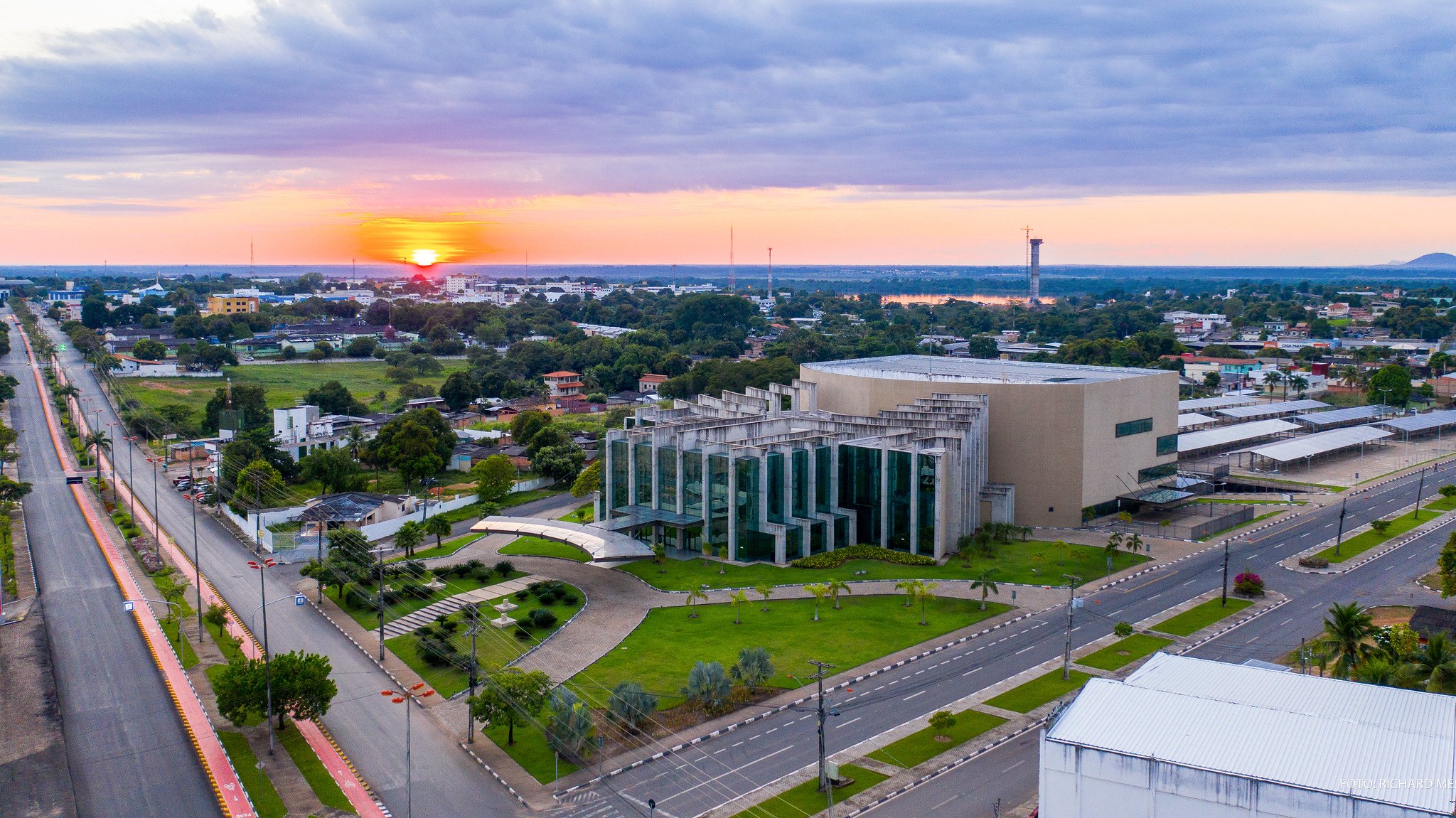 Aerial view of a mid-sized Brazilian city at sunset, with a modernist glass-and-concrete civic building surrounded by green lawns, wide avenues and a glowing orange horizon — the gateway city for the Mount Roraima trek