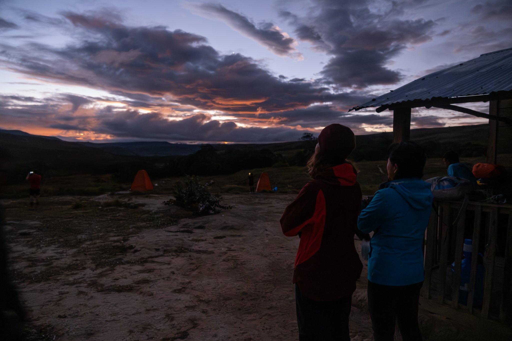 Trekkers in warm jackets watch a dramatic orange and purple sunset from a hillside campsite during a Mount Roraima trek, with glowing orange tents visible in the background