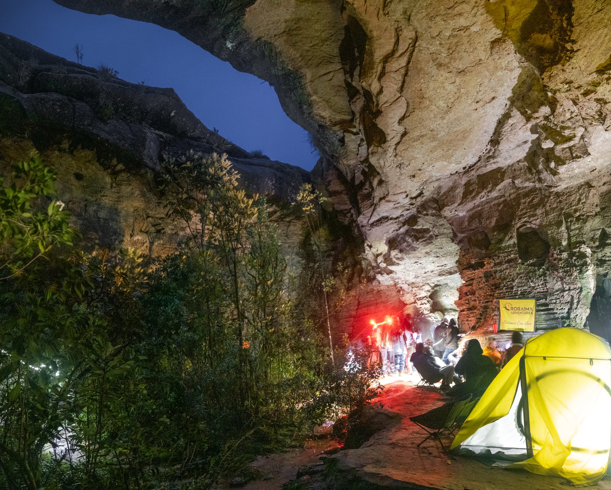 Trekkers gathered around a glowing campfire inside a vast sandstone rock shelter at night during a Mount Roraima trek, a yellow tent lit from within in the foreground and a Roraima Adventures banner visible on the cave wall