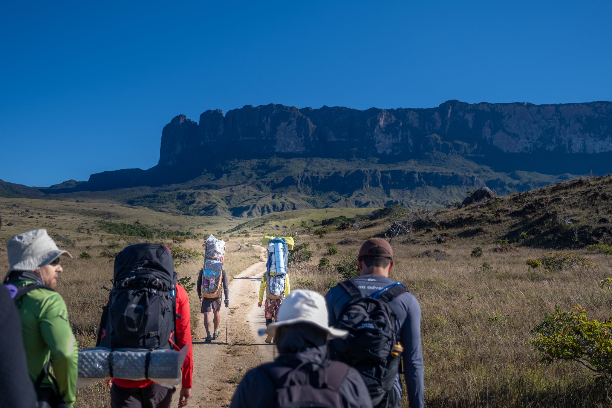 A group of backpackers on the Mount Roraima trek walking a dirt trail toward the sheer sandstone cliffs of the tepui under a deep blue sky