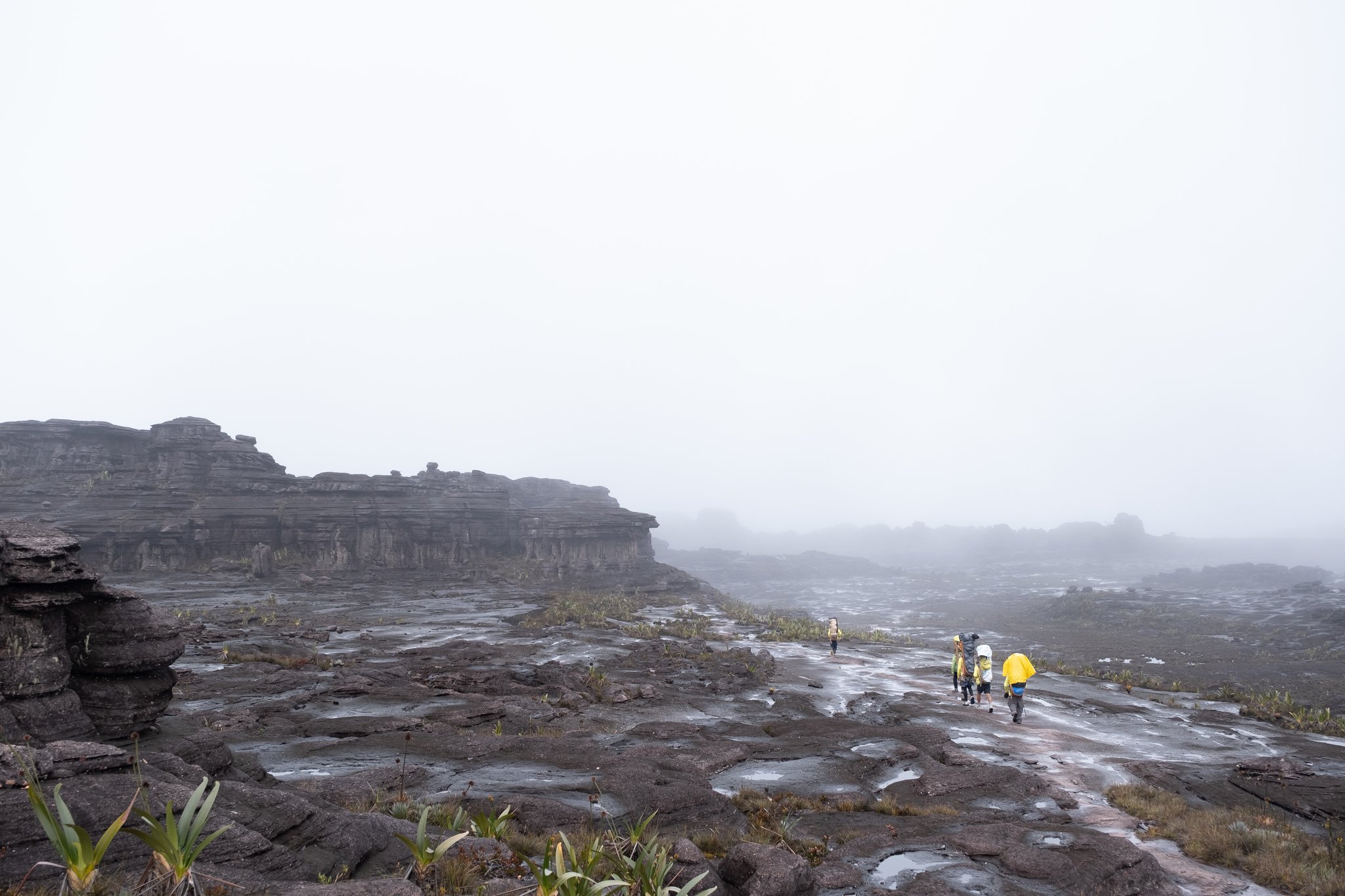A small group of trekkers in yellow rain gear crosses a vast, rain-soaked rocky plateau on the Mount Roraima trek, with ancient sandstone cliffs looming through thick mist
