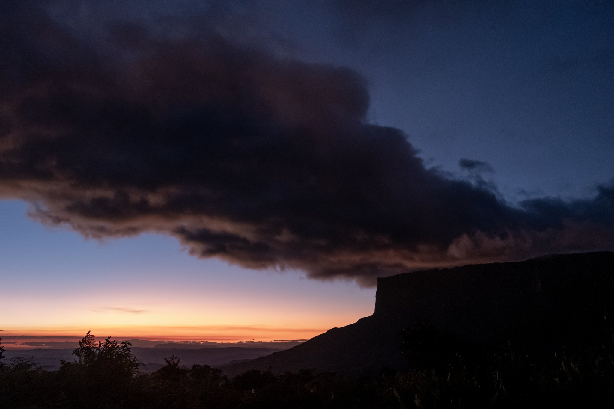 A dramatic storm cloud spreads over the silhouetted cliff edge of a tepui during a fiery Mount Roraima trek sunset, with an orange and deep-blue sky on the horizon