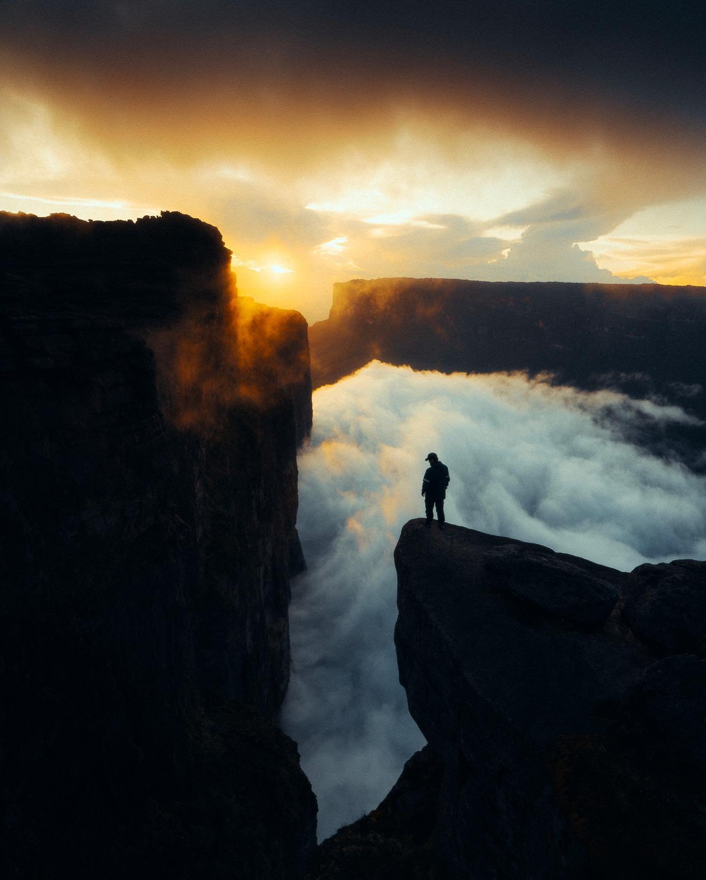 A lone hiker silhouetted on a rocky cliff edge during a Mount Roraima trek, with a sea of clouds filling the canyon below and a golden sunset blazing between sheer tepui walls