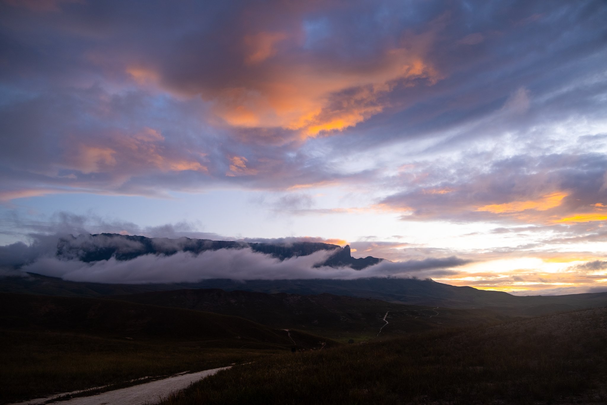A dramatic sunset sky of orange and purple clouds hovers over a flat-topped tepui silhouette shrouded in low cloud during a Mount Roraima trek, with a winding trail visible across the dark savannah foreground