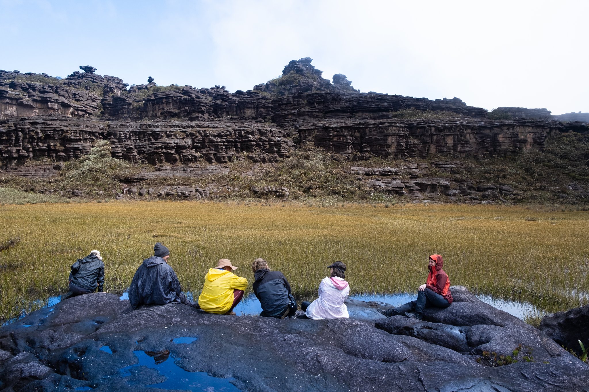 Six trekkers in rain gear sit on dark wet rocks overlooking a golden grassland meadow enclosed by ancient layered sandstone cliffs on the Mount Roraima trek
