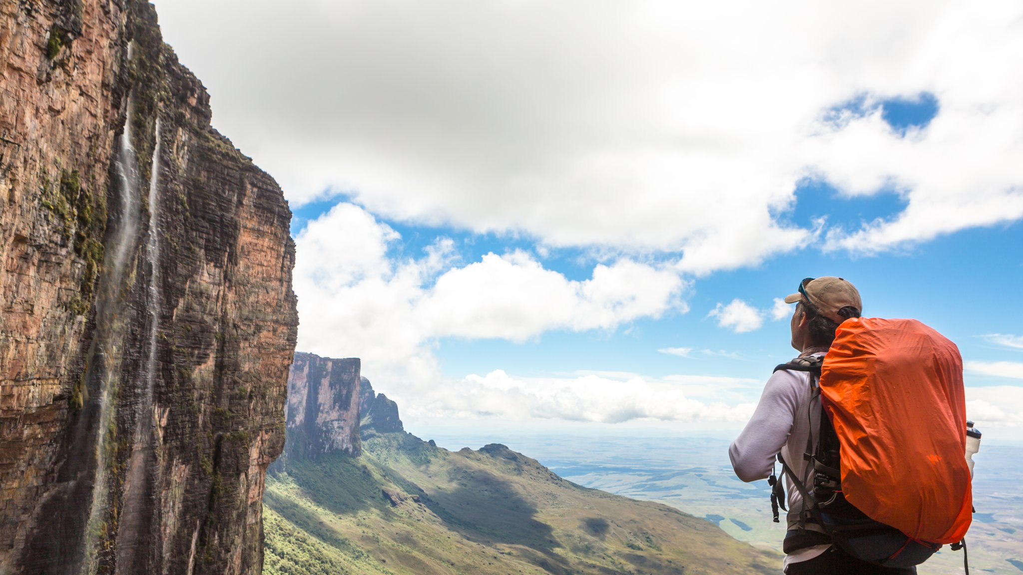 A hiker with an orange-covered backpack stands at the edge of a towering sandstone cliff face on the Mount Roraima trek, gazing across a vast savannah landscape as a thin waterfall cascades down the sheer vertical wall beside them
