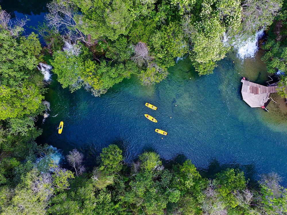 Aerial view of four yellow kayaks paddling along a crystal-clear turquoise river flanked by dense green forest on a Pantanal Bonito tour, with a wooden dock visible on the bank