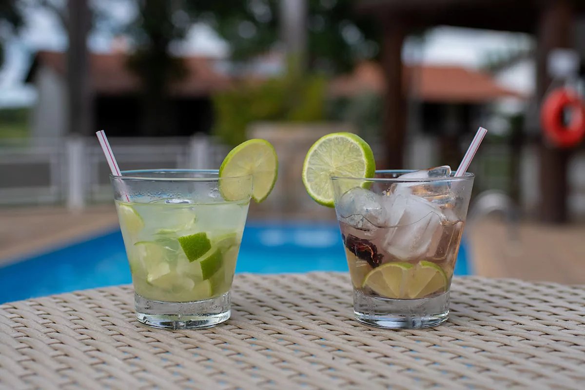 Cold drinks, warm air, and nowhere else to be. Two lime-garnished cocktails on a wicker table beside a swimming pool, served during a Pantanal Bonito tour lodge stay