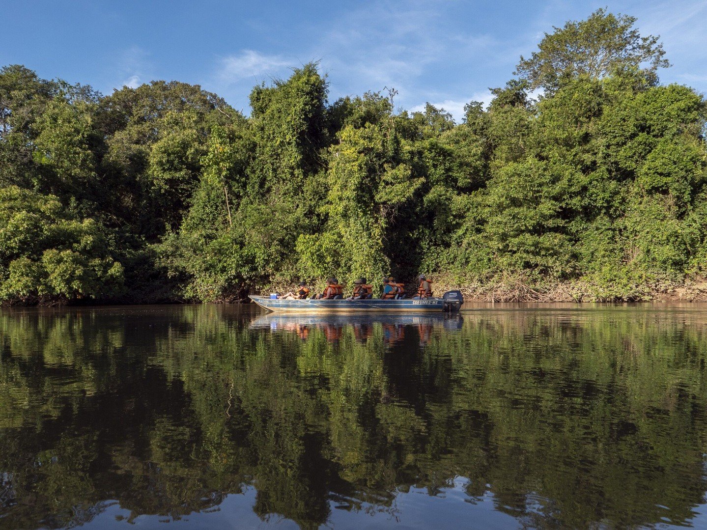 Still water, towering trees, and a boatload of wide-eyed explorers. A small motorboat carrying wildlife watchers in orange life vests glides along a calm river channel lined with dense green riparian forest on a Pantanal Bonito tour