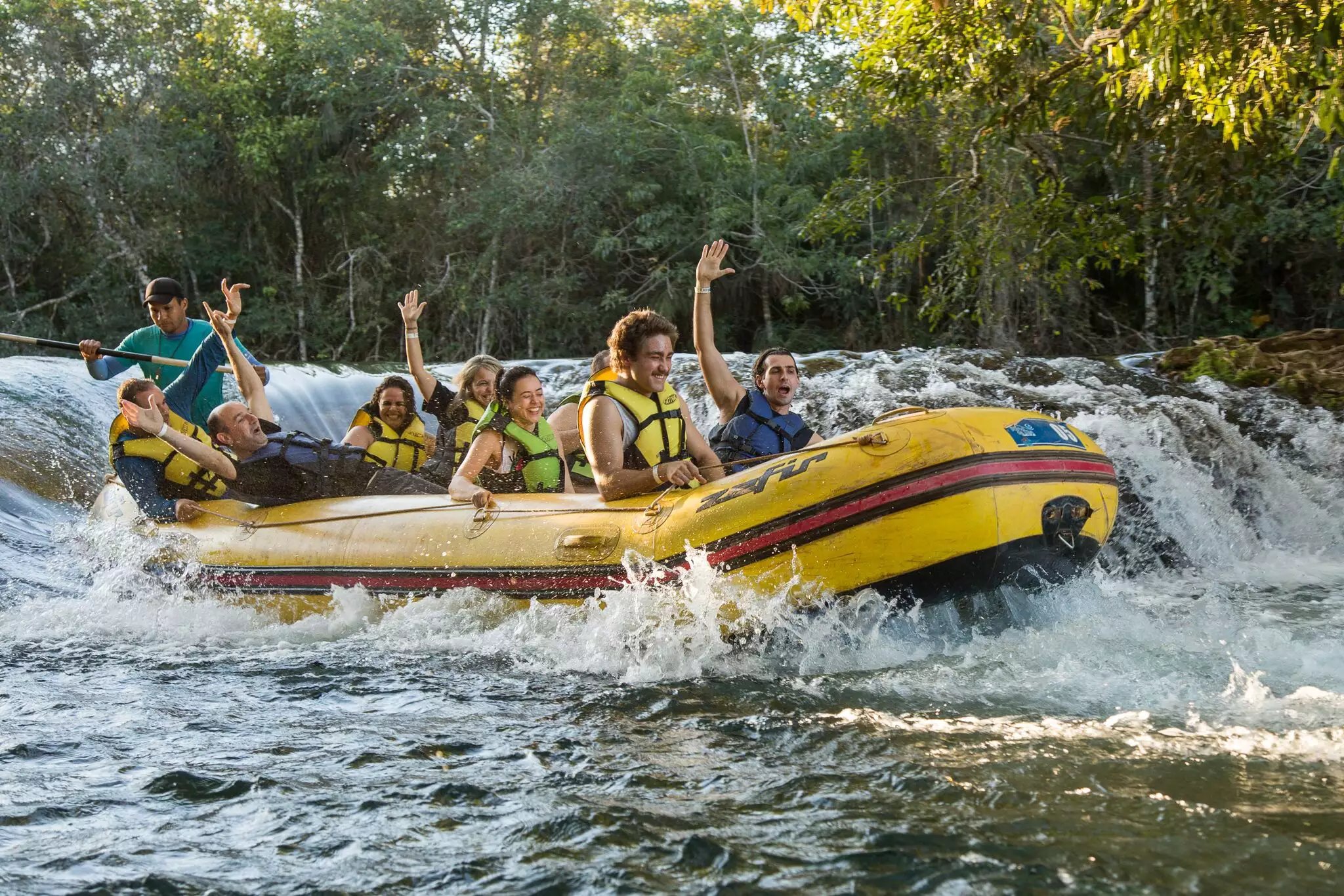 Hands up, rapids ahead — pure adrenaline on the river. A group of cheering guests in life jackets ride a yellow inflatable raft through churning white-water rapids on a Pantanal Bonito tour, surrounded by lush riverside forest
