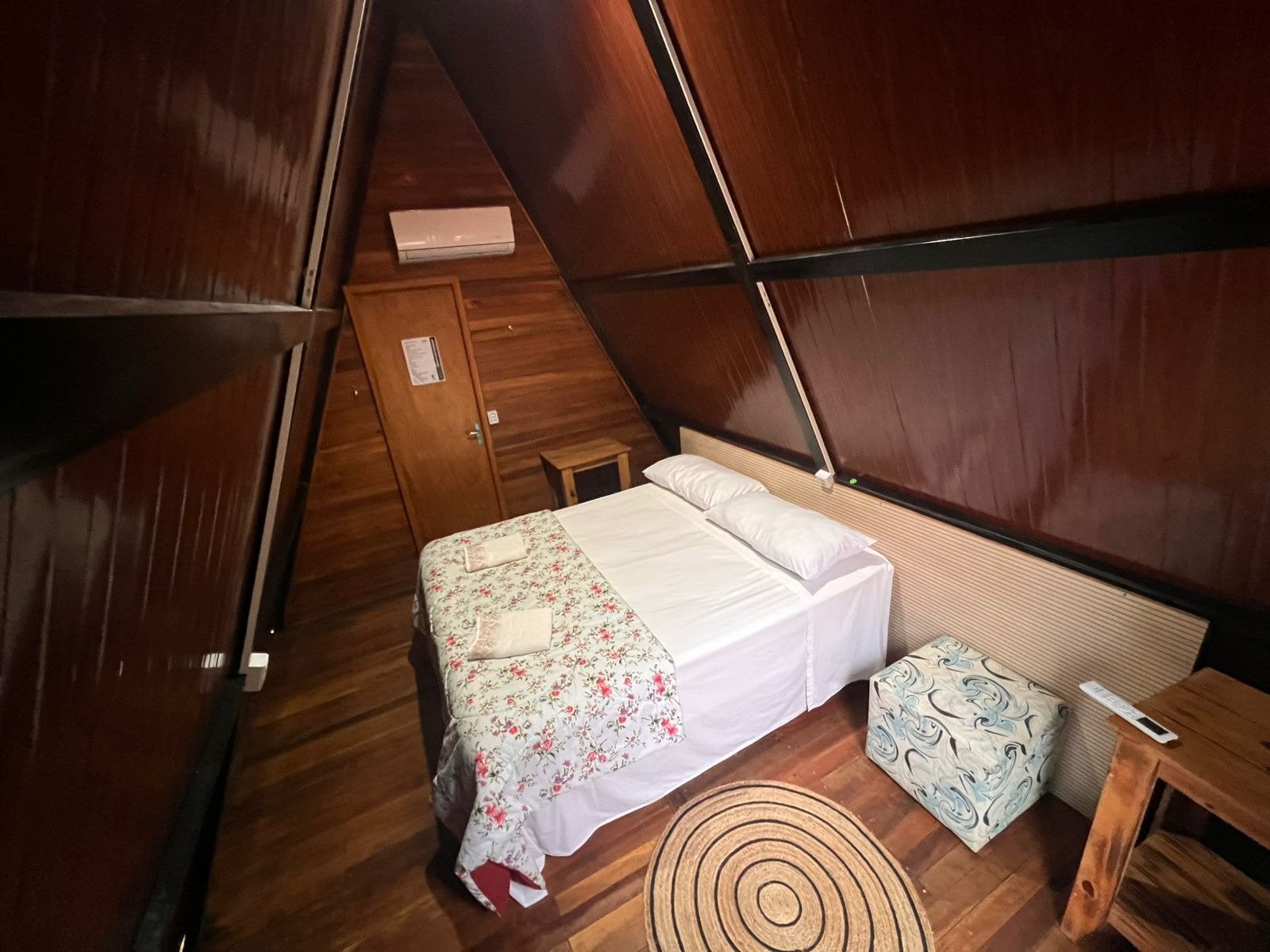 Simple, clean, and cosy — your base between jaguar sightings. Interior of an A-frame wooden cabin with a double bed, floral bedspread, timber floors, and air conditioning, offered on a pantanal jaguar budget safari