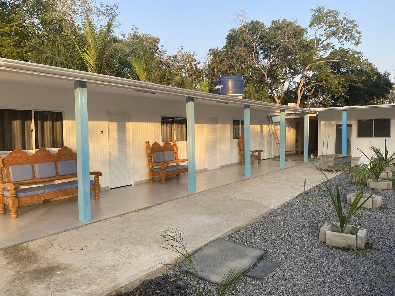 Carved benches waiting on the verandah as the afternoon light fades. A row of numbered guest rooms with carved wooden benches on a covered verandah, surrounded by tropical trees — simple, comfortable accommodation on a pantanal jaguar budget safari
