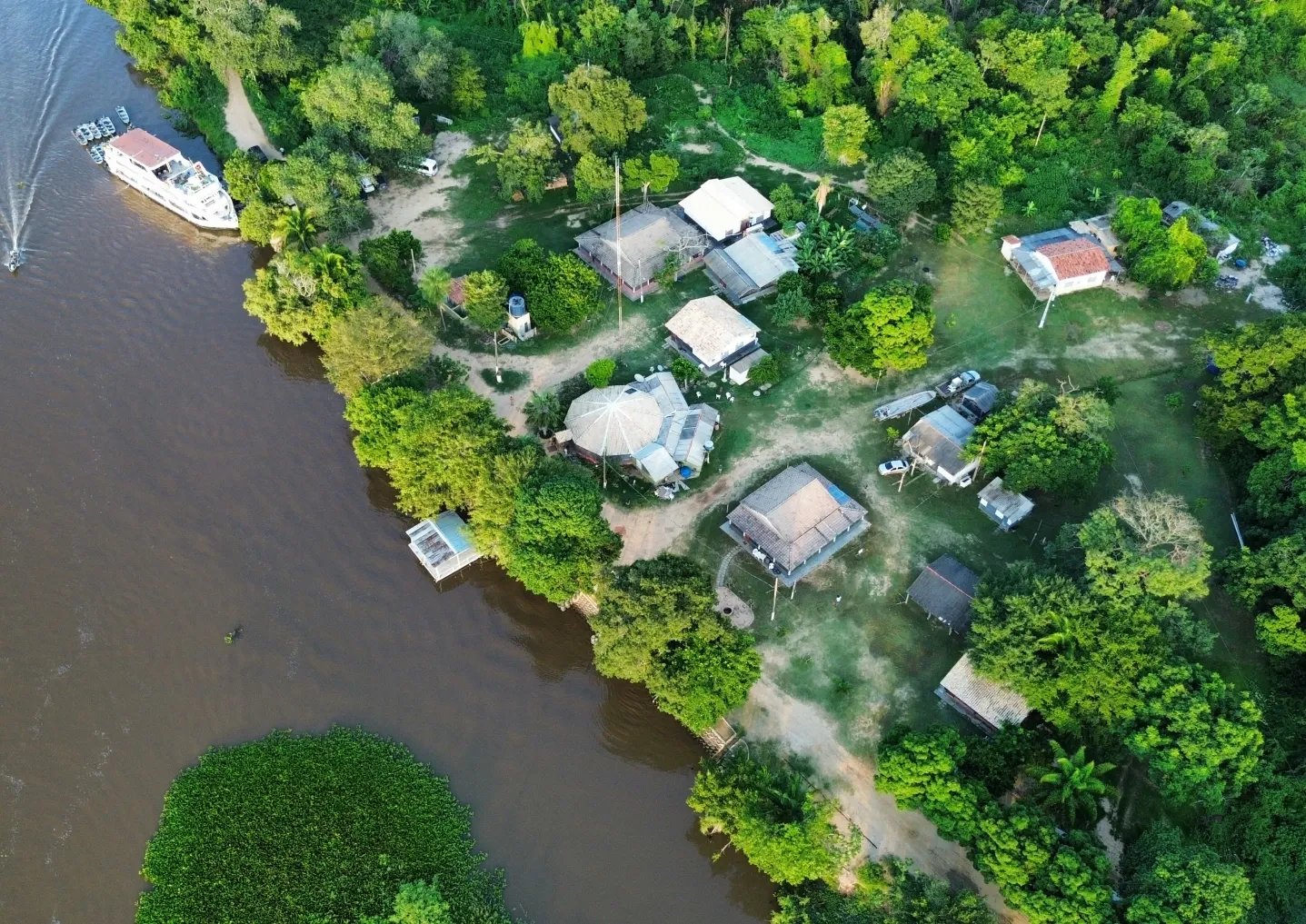 Jungle canopy on three sides, a brown river on the fourth — your base for a Pantanal jaguar budget safari. Aerial drone view of a small riverside lodge tucked into dense green forest along a brown river in the Pantanal, with a multi-deck boat moored at the bank and thatched-roof buildings set among tropical trees