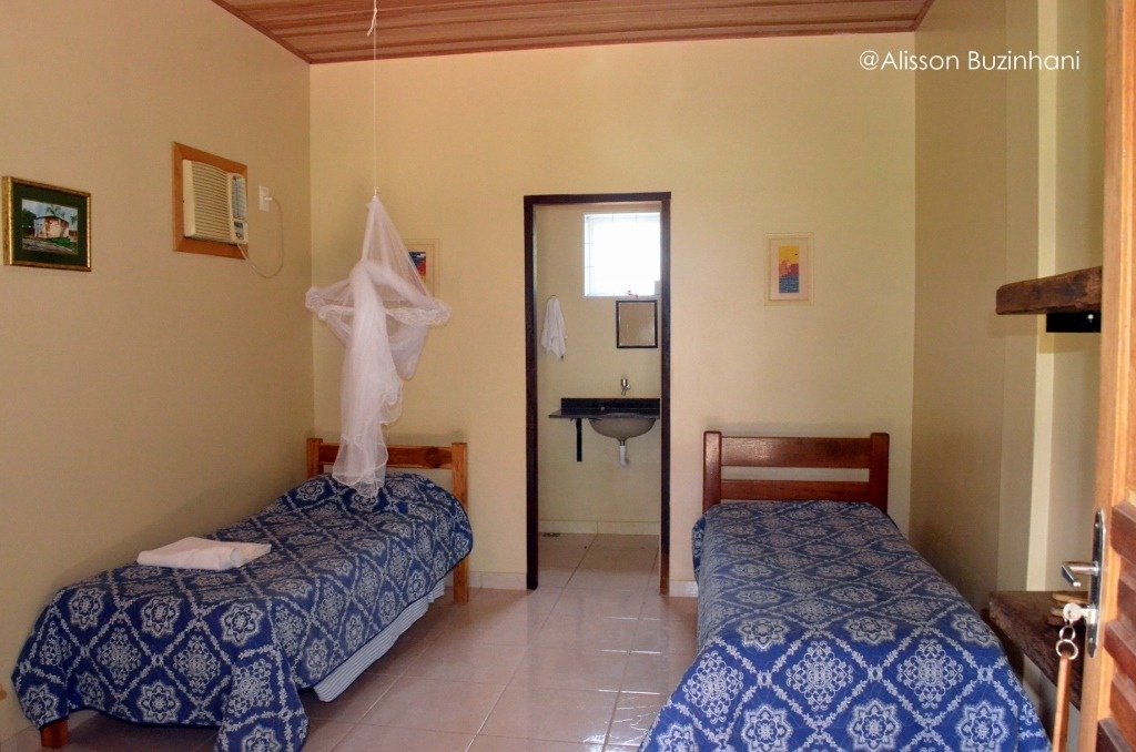 Mosquito net overhead, matching beds ready — simple comfort after a long day on the river. Twin bedroom with blue patterned bedspreads, a hanging mosquito net, and an en-suite bathroom visible through the doorway at a simple Pantanal jaguar budget safari lodge