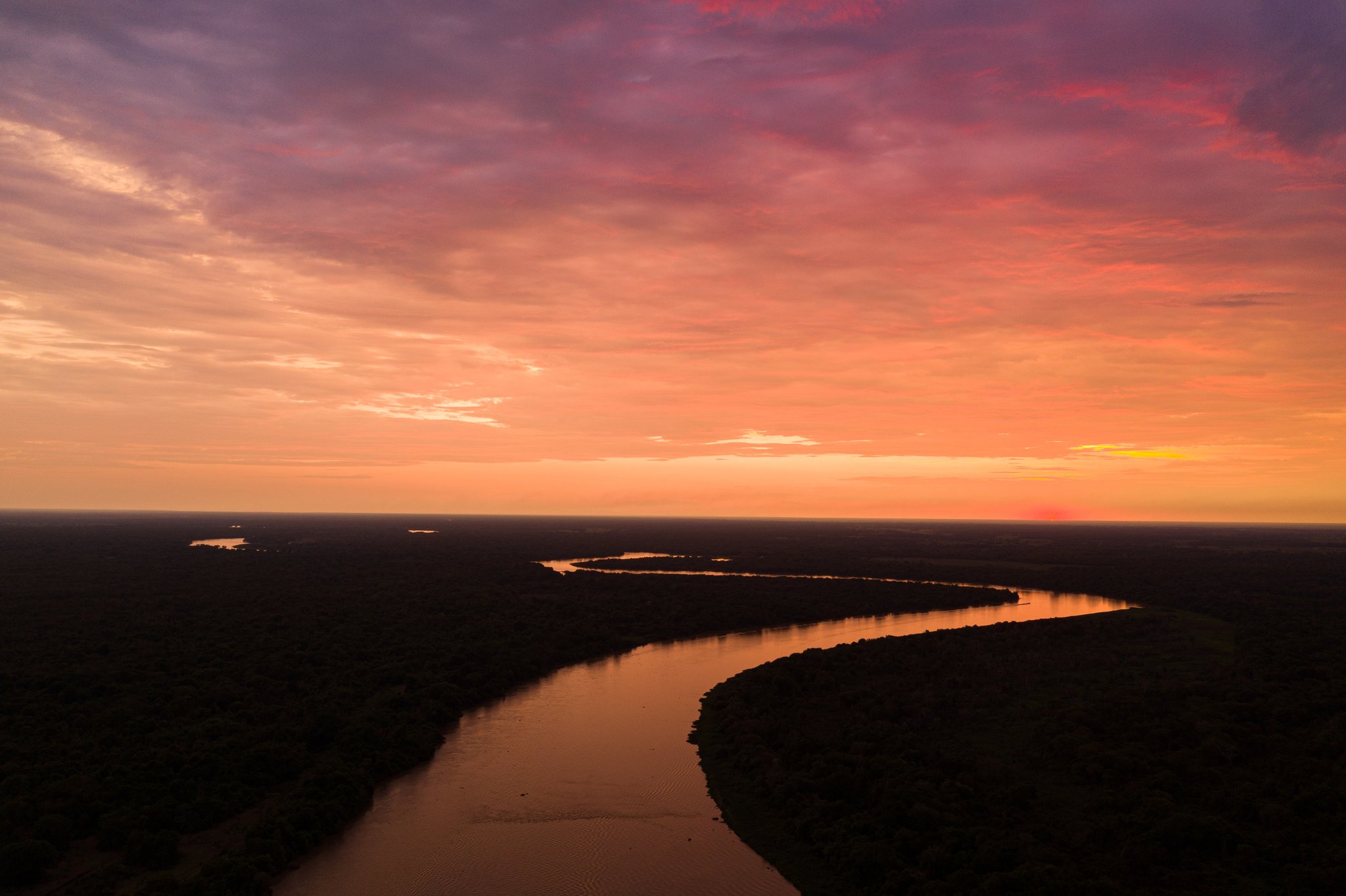 Aerial view of a winding river cutting through the dark Pantanal wetlands at sunset, with a vivid orange, pink and purple sky reflected on the water — a landscape at the heart of pantanal jaguar tours