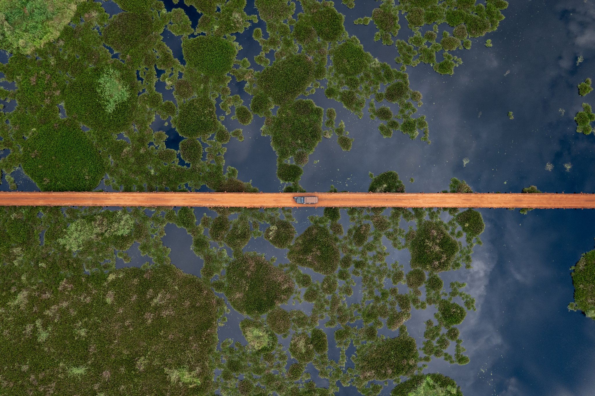 Aerial top-down view of a red dirt road crossing a vast flooded wetland dotted with green vegetation islands, with a single vehicle tiny at the centre — a striking scale of the pantanal luxury safari landscape