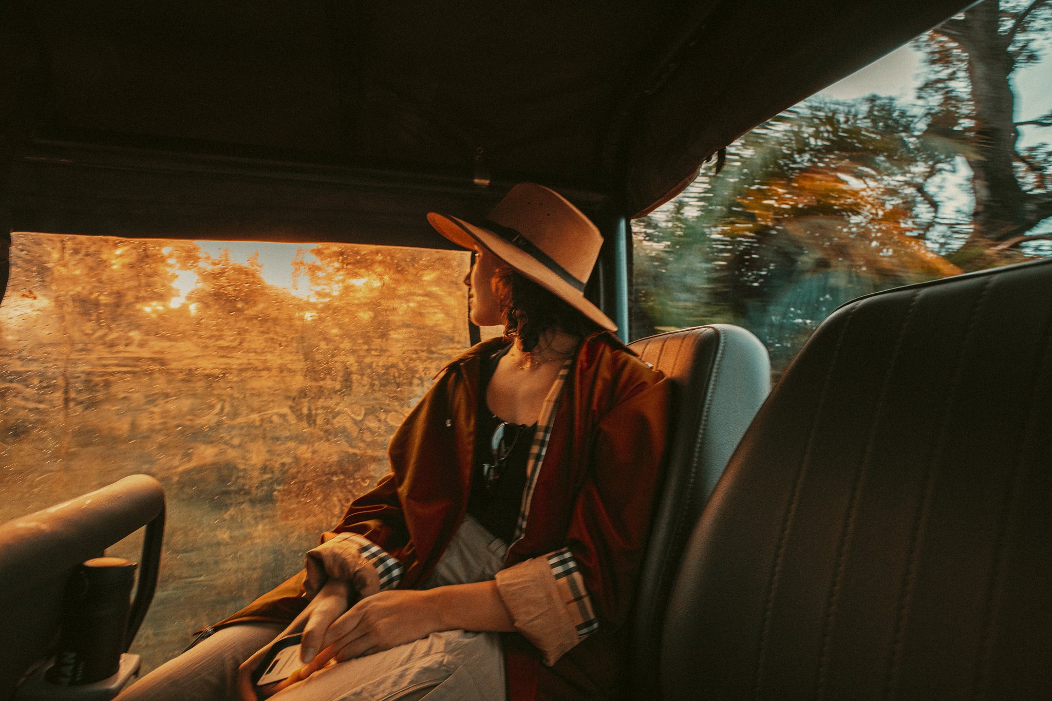 Sunset light catches the rain on the glass — the wild already beginning. Photo: Felipe Castellari A traveller in a wide-brim hat and rust-coloured wrap gazes out a rain-speckled jeep window at golden-hour light filtering through trees during a pantanal luxury safari transfer