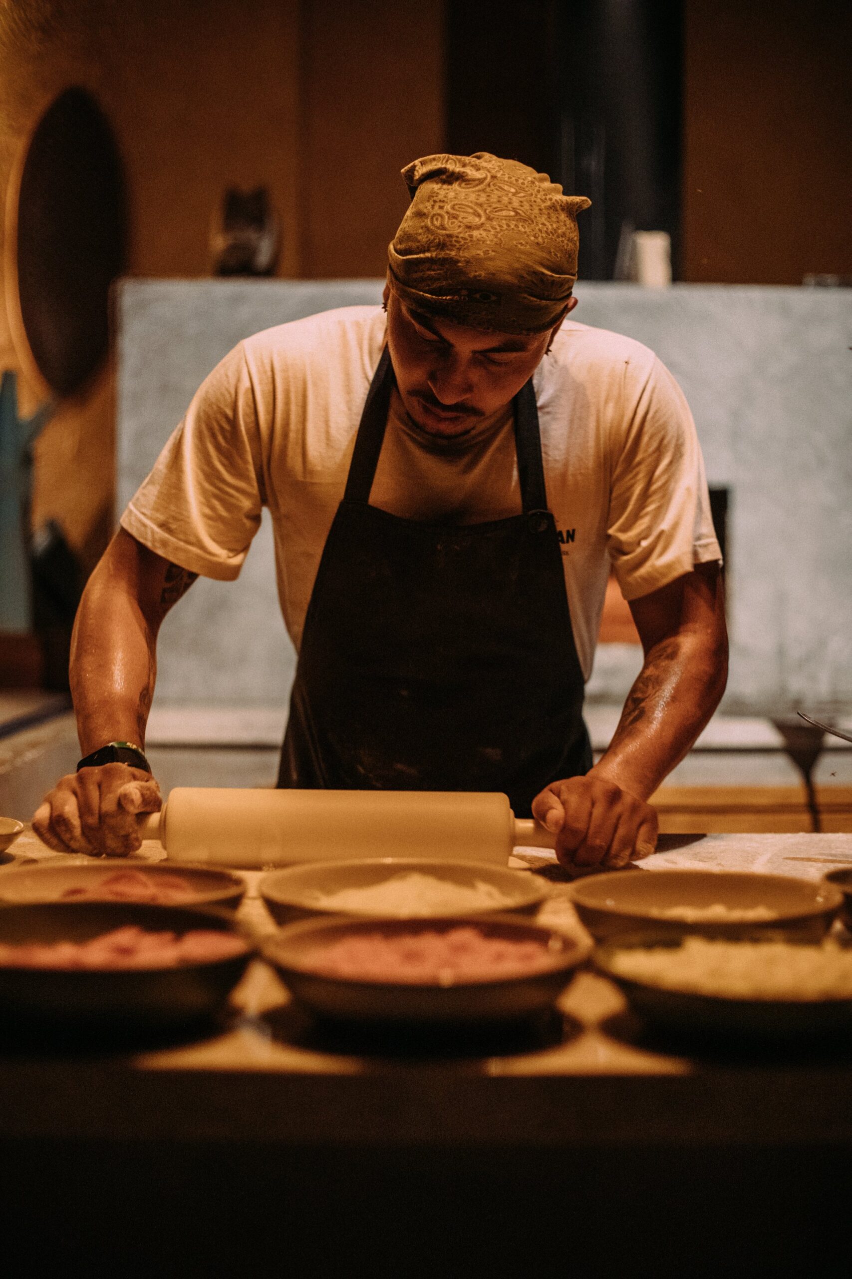 Focused, unhurried craft — fresh pastry prepared by hand before every service. A chef wearing a dark apron and bandana rolls out dough at a wooden prep station lined with ceramic bowls of filling, part of the pantanal luxury safari dining experience at Caiman