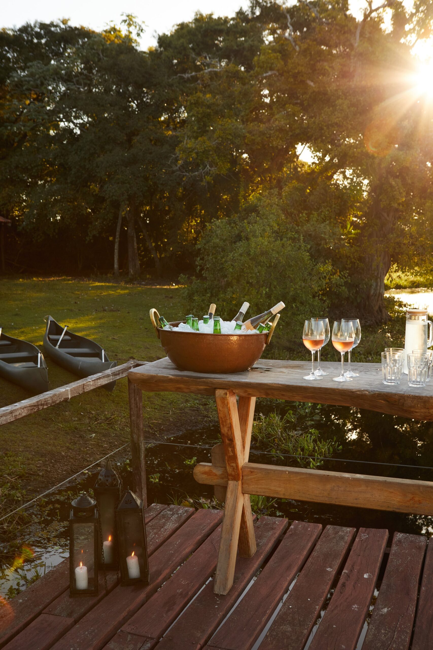 Chilled rosé, candlelight, canoes at rest — the evening ritual before dinner. A copper ice bucket filled with chilled bottles and rosé wine glasses on a rustic wooden table at sunset on a riverside deck, with canoes on the bank and candlelit lanterns below — part of the pantanal luxury safari experience at Caiman
