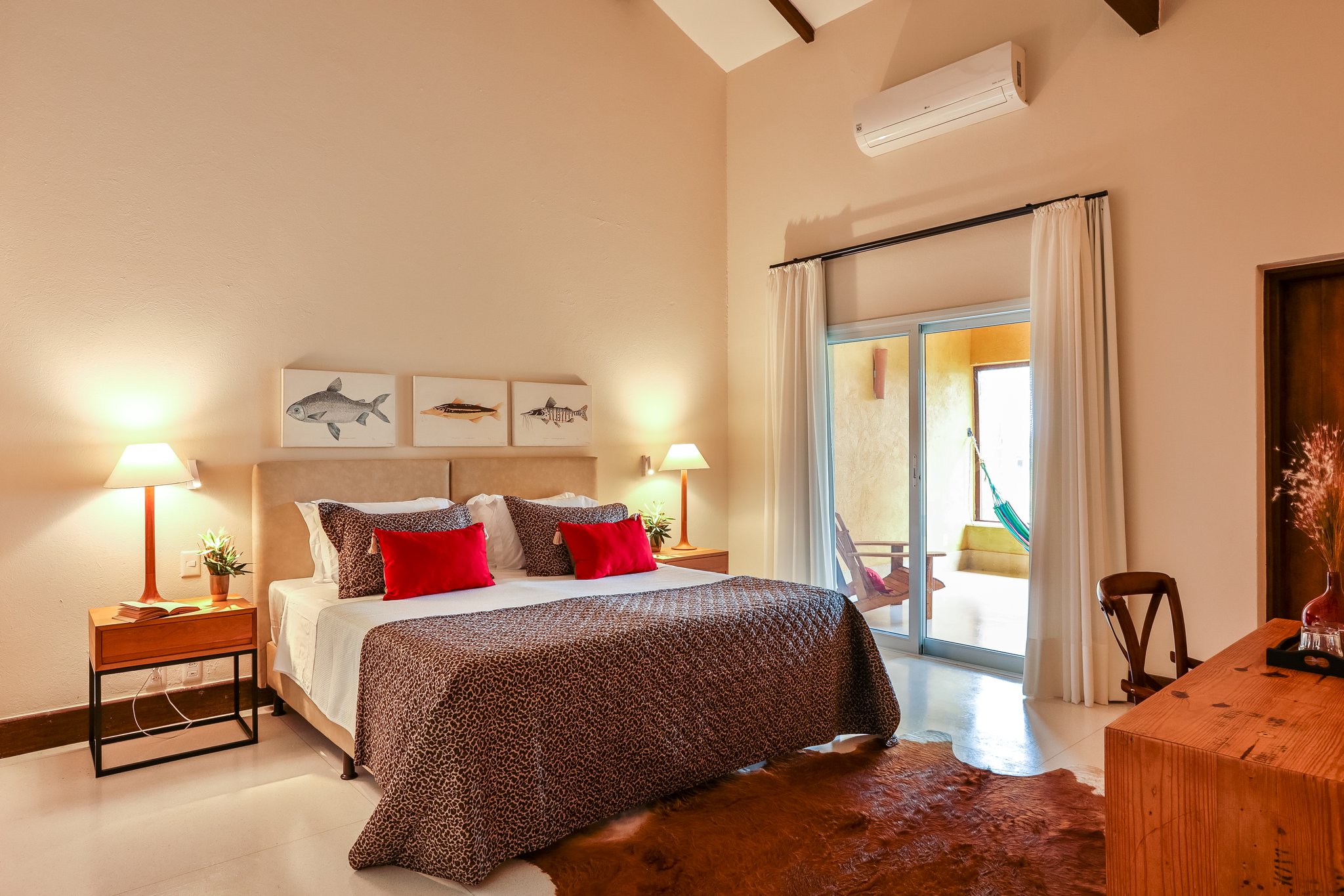 Crisp white linens, warm lamplight, and a hammock waiting just beyond the glass — rest comes easy after a day in the wild. Spacious pantanal luxury safari suite with a king bed dressed in leopard-print throws and red cushions, fish artwork above the headboard, vaulted timber ceiling, and a private terrace with a hammock visible through sliding glass doors