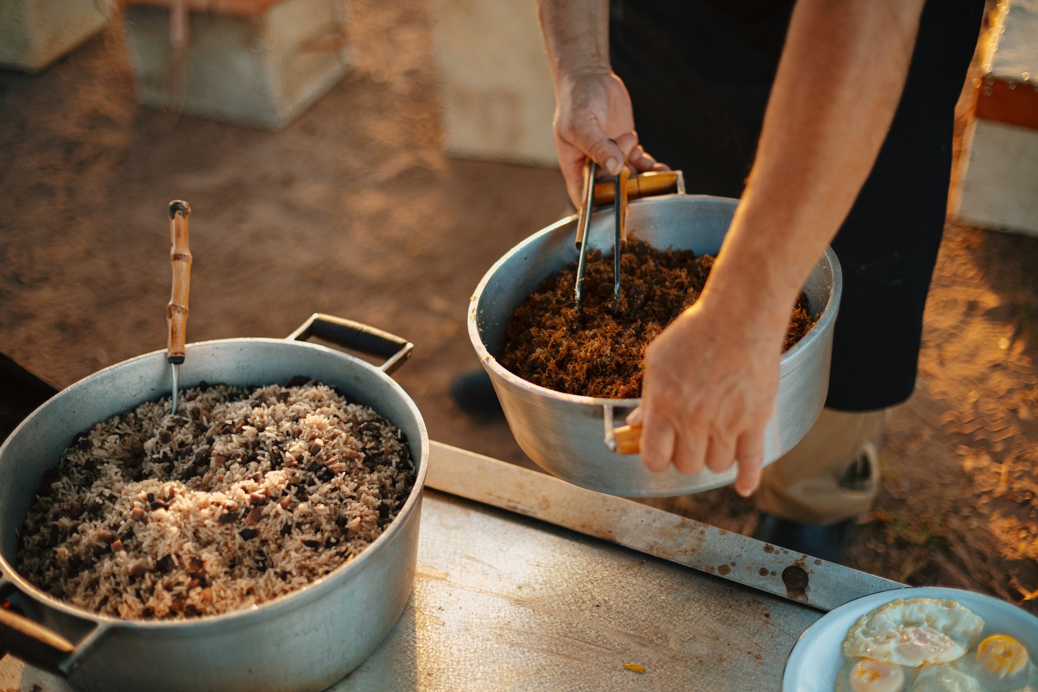 Generous pots of rice, beans, and spiced meat — fuel for a full day in the wild. Hands stirring seasoned shredded meat in an aluminium pot beside a large pot of rice and black beans, served outdoors as part of a pantanal luxury safari all-inclusive meal