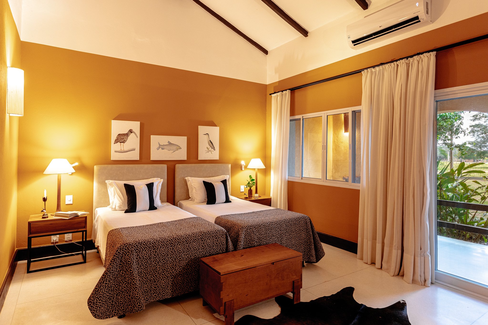 Safari-inspired décor and a private garden view — rest comes easy between expeditions. Photo: Felipe Castellari Twin bedroom at a pantanal luxury safari lodge featuring warm ochre walls, wildlife botanical prints, leopard-print bedspreads, exposed timber ceiling beams, and a sliding glass door opening onto lush greenery