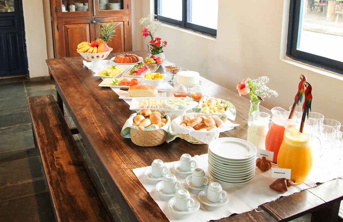 A generous Brazilian breakfast buffet laid out on a long wooden table at a Pantanal tour from Campo Grande lodge, featuring fresh tropical fruits, bread rolls, cakes, acerola and maracujá juices, and small espresso cups
